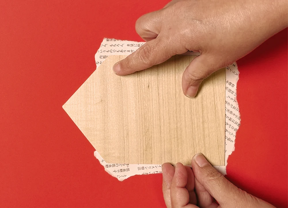 Mãos colando um recorte de madeira sobre papel vermelho, parte do processo de colagem