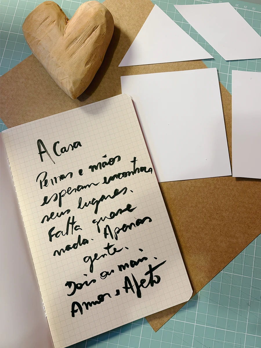 Caderno aberto com o poema manuscrito 'A Casa', ao lado de um coração de madeira esculpido e recortes de papel kraft